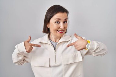 Orta yaşlı İspanyol bir kadın izole bir arka planda duruyor. Yüzünde bir gülümsemeyle, gururlu ve mutlu bir şekilde kendini işaret ediyor.. 