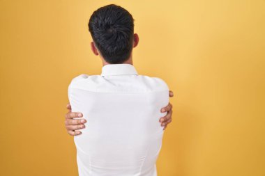 Sarı arka planda dikilen İspanyol genç adam kendine sarılıyor mutlu ve pozitif bir şekilde. Sevgi ve özgüven. 