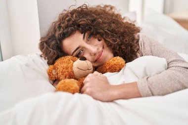 Young hispanic woman lying on bed hugging teddy bear at bedroom