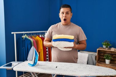 Hispanic young man holding folded laundry after ironing afraid and shocked with surprise and amazed expression, fear and excited face. 