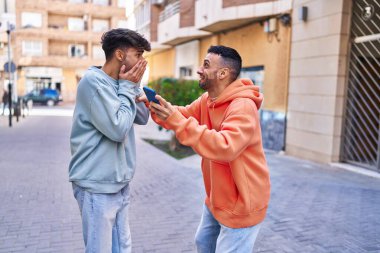 İki erkek caddede birbirlerine akıllı telefon kullanarak sarılıyorlar.