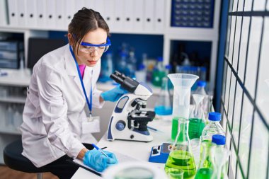 Young beautiful hispanic woman scientist using microscope writing on document at laboratory