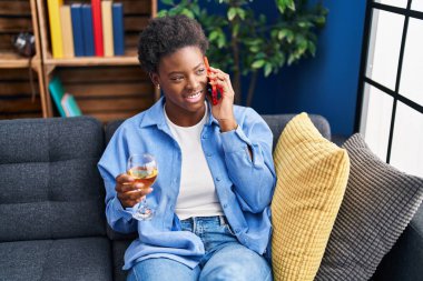 Afro-Amerikalı kadın akıllı telefondan konuşuyor evde şampanya içiyor.