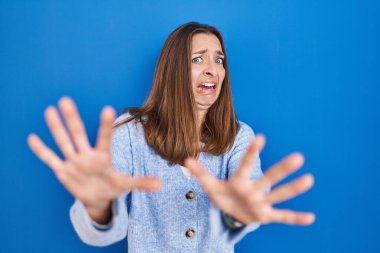 Young woman standing over blue background afraid and terrified with fear expression stop gesture with hands, shouting in shock. panic concept. 