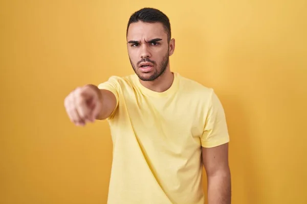 カメラに不満と不満を示す黄色の背景の上に立っている若いヒスパニック系の男 怒っているとあなたと激怒 — ストック写真