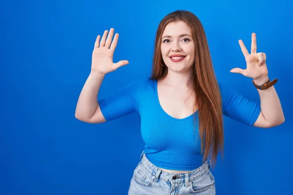 自信と幸せ笑いながら指の数8で見せ 指差す青い背景に立って赤毛の女性 — ストック写真