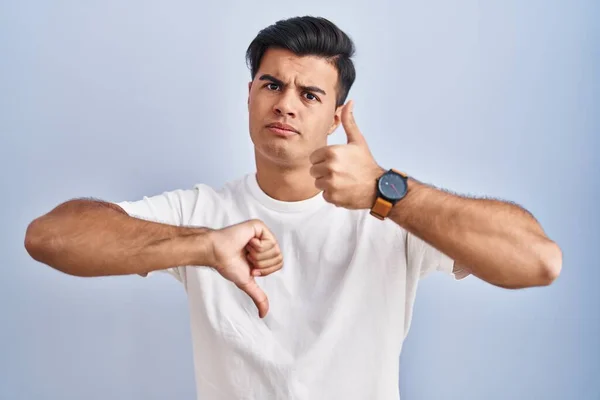 Hispanic man standing over blue background doing thumbs up and down, disagreement and agreement expression. crazy conflict