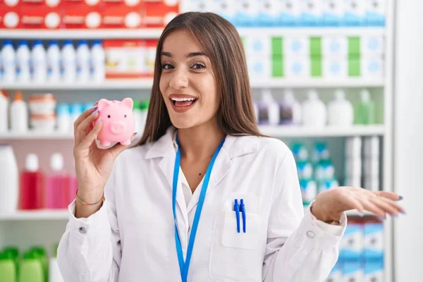 薬局で働く若いブルネットの女性は幸せな笑顔で達成を祝う貯金箱を保持し 手を上げと勝者の式 — ストック写真