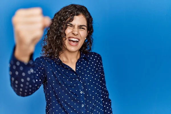 Young brunette woman with curly hair wearing casual clothes over blue background angry and mad raising fist frustrated and furious while shouting with anger. rage and aggressive concept.
