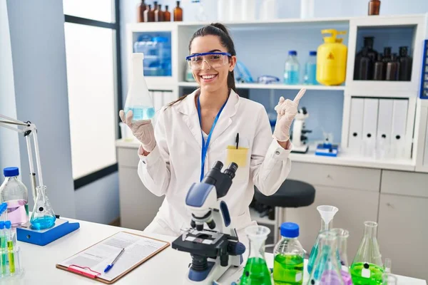 Hispanic woman working at scientist laboratory smiling happy pointing with hand and finger to the side