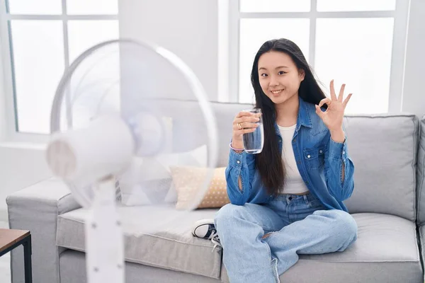 stock image Young asian woman drinking glass of water enjoying air from fan doing ok sign with fingers, smiling friendly gesturing excellent symbol 