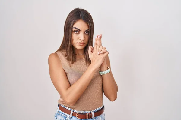 Young hispanic woman standing over white background holding symbolic gun with hand gesture, playing killing shooting weapons, angry face