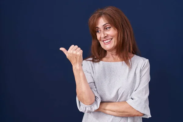 Middle age woman standing over blue background smiling with happy face looking and pointing to the side with thumb up.