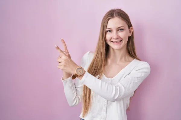 年轻的高加索女人笑着站在粉色的背景上 看着摄像机用双手和手指指向旁边 — 图库照片
