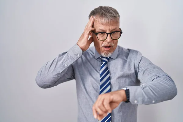 Hispanic business man with grey hair wearing glasses looking at the watch time worried, afraid of getting late