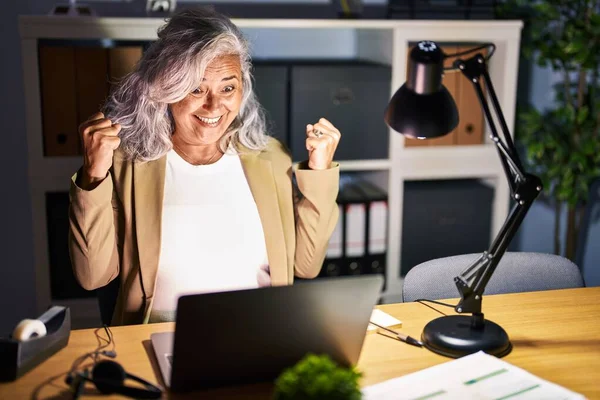 深夜にコンピュータのラップトップを使用して灰色の髪の仕事をしている中年の女性は非常に満足し 腕を上げて勝者のジェスチャーを行う興奮し 笑顔と成功のために叫んでいます お祝いのコンセプト — ストック写真