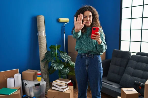 Young hispanic woman at new home using smartphone with open hand doing stop sign with serious and confident expression, defense gesture