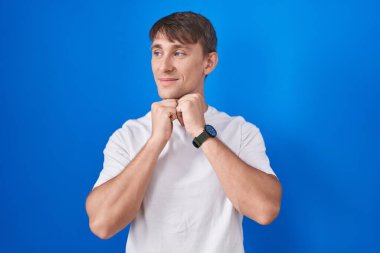 Mavi arka planda dikilen beyaz tenli sarışın adam. Gergin ve heyecanlı bir şekilde çenesinin kenarına bakıyor. 