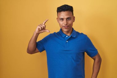 Young hispanic man standing over yellow background smiling and confident gesturing with hand doing small size sign with fingers looking and the camera. measure concept. 