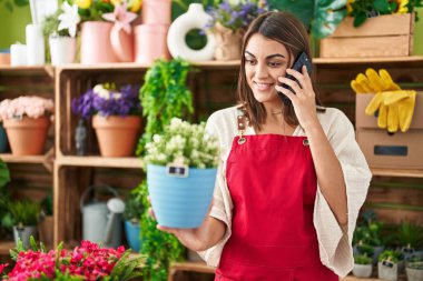 Young beautiful hispanic woman florist talking on smartphone holding plant at flower shop