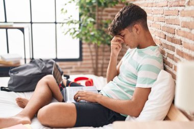 Young hispanic teenager student stressed using laptop sitting on bed at bedroom