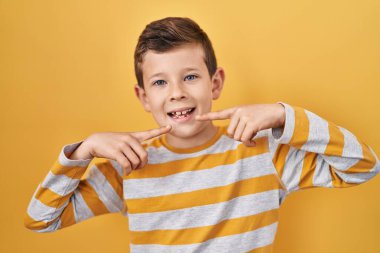 Sarı arka planda duran, neşeli gülümseyen, parmaklarıyla ve ağzıyla işaret eden beyaz bir çocuk. diş sağlığı kavramı. 