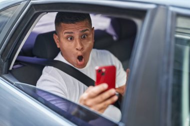 Young hispanic man doing video call with smartphone in the car scared and amazed with open mouth for surprise, disbelief face 