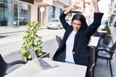 Genç ve güzel İspanyol kadın, kahve dükkanının terasındaki masada oturan ve kazanan yüz ifadesiyle dizüstü bilgisayar kullanıyor.