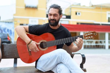 Young hispanic man musician playing classical guitar sitting on bench at street