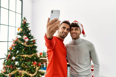 İki İspanyol erkek akıllı telefonun yanında selfie çekiyor. Evde Noel ağacının yanında duruyorlar.