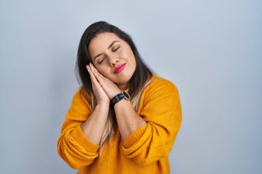 Young hispanic woman standing over isolated background sleeping tired dreaming and posing with hands together while smiling with closed eyes. 