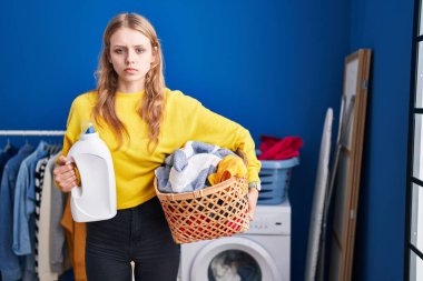 Young caucasian woman holding laundry basket and detergent bottle skeptic and nervous, frowning upset because of problem. negative person. 
