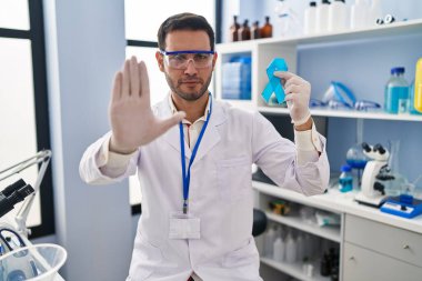 Young hispanic man with beard working at scientist laboratory holding blue ribbon with open hand doing stop sign with serious and confident expression, defense gesture 