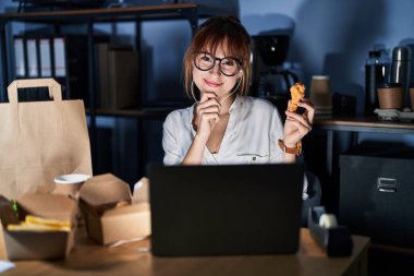 Genç ve güzel bir kadın bilgisayar laptopu kullanıyor ve yemek yiyor kameraya kendinden emin bakıyor kollarını kavuşturmuş ve çenesini kaldırmış gülümsüyor. Olumlu düşünmek. 