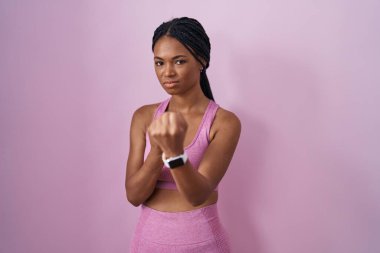 Pembe arka planda spor kıyafetleri giyen Afro-Amerikalı bir kadın. Yumruk savunma hareketleriyle dövüşmeye hazır. Kızgın ve üzgün. 
