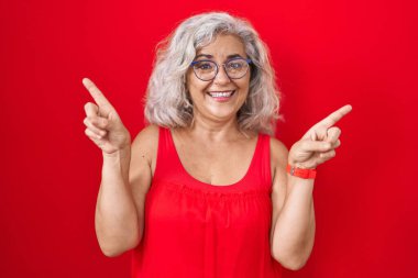 Middle age woman with grey hair standing over red background smiling confident pointing with fingers to different directions. copy space for advertisement 