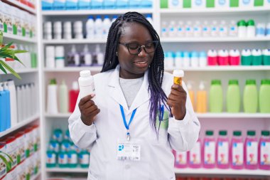 African american woman pharmacist smiling confident holding pills bottles at pharmacy