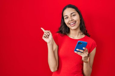 Genç Brezilyalı kadın kırmızı arka planda akıllı telefon kullanıyor. Yüzünde kocaman bir gülümseme var. Elleriyle kameraya bakıyor.. 