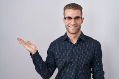 Young caucasian man standing over isolated background smiling cheerful presenting and pointing with palm of hand looking at the camera. 