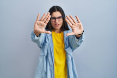 Mavi arka planda dikilen Latin kökenli genç bir kadın avuçlarıyla jestleri durduruyor. Öfkeli ve hayal kırıklığına uğramış bir ifade. 
