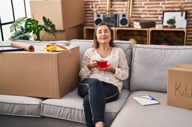 Middle age woman drinking coffee sitting on sofa at new home