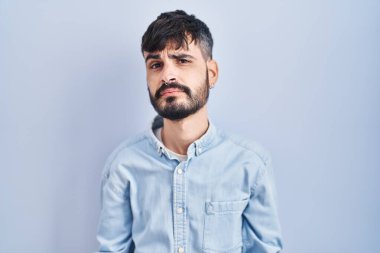 Young hispanic man with beard standing over blue background clueless and confused with open arms, no idea concept. 