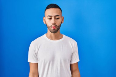 Young hispanic man standing over blue background making fish face with lips, crazy and comical gesture. funny expression. 