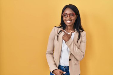 Yüzünde mutlu ve doğal bir ifadeyle elleri ve parmaklarıyla işaret eden neşeli yüzlü, gözlüklü Afrikalı genç bir kadın. 