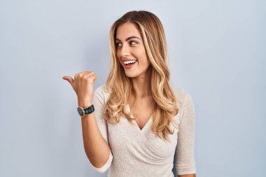 Young blonde woman standing over isolated background smiling with happy face looking and pointing to the side with thumb up. 