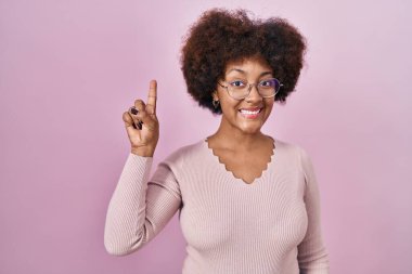 Young african american woman standing over pink background showing and pointing up with finger number one while smiling confident and happy. 