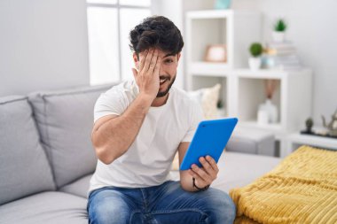 Hispanic man with beard using touchpad sitting on the sofa covering one eye with hand, confident smile on face and surprise emotion. 