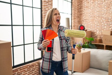 Young blonde woman holding roller painter and paint samples at new home angry and mad screaming frustrated and furious, shouting with anger. rage and aggressive concept. 
