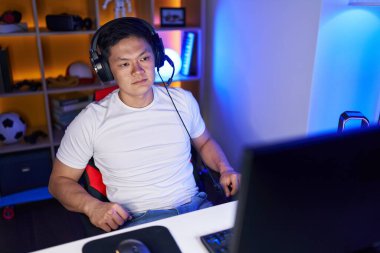 Young chinese man streamer sitting on table with serious expression at gaming room