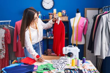 Young beautiful hispanic woman tailor holding t shirt writing on notebook at clothing factory